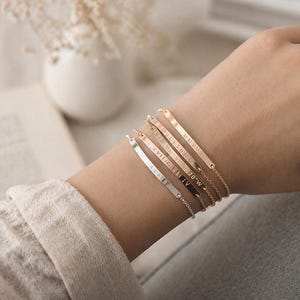 May include: Several delicate bar bracelets in gold and silver hues, each engraved with custom text. The bracelets are shown on a wrist, highlighting their fine chain details and personalised inscriptions.