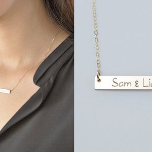 Puede incluir: Collar de barra dorado con los nombres "Sam & Liam" grabados en un colgante rectangular. El collar se muestra en el cuello de una persona, con un primer plano del colgante y la cadena.