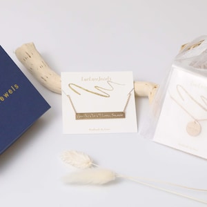 Puede incluir: Un surtido de joyas de LuvLace Jewels. Una caja azul marino con letras doradas se encuentra junto a dos collares en tarjetas y un collar en una bolsa transparente. Un collar tiene un colgante de barra con texto, el otro tiene un colgante redondo.