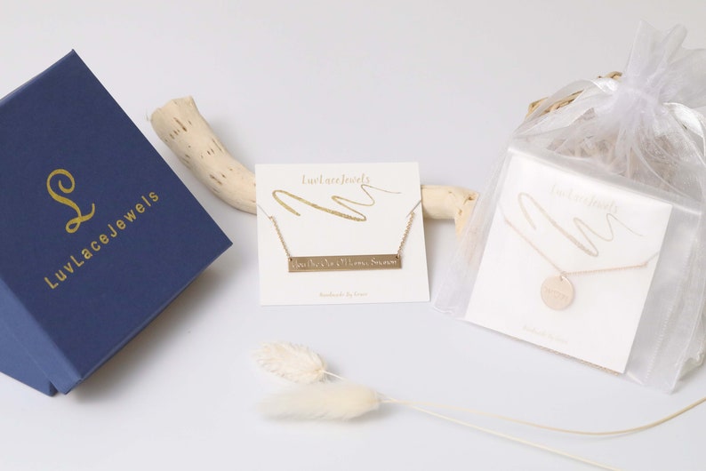 Puede incluir: Un surtido de joyas de LuvLace Jewels. Una caja azul marino con letras doradas se encuentra junto a dos collares en tarjetas y un collar en una bolsa transparente. Un collar tiene un colgante de barra con texto, el otro tiene un colgante redondo.