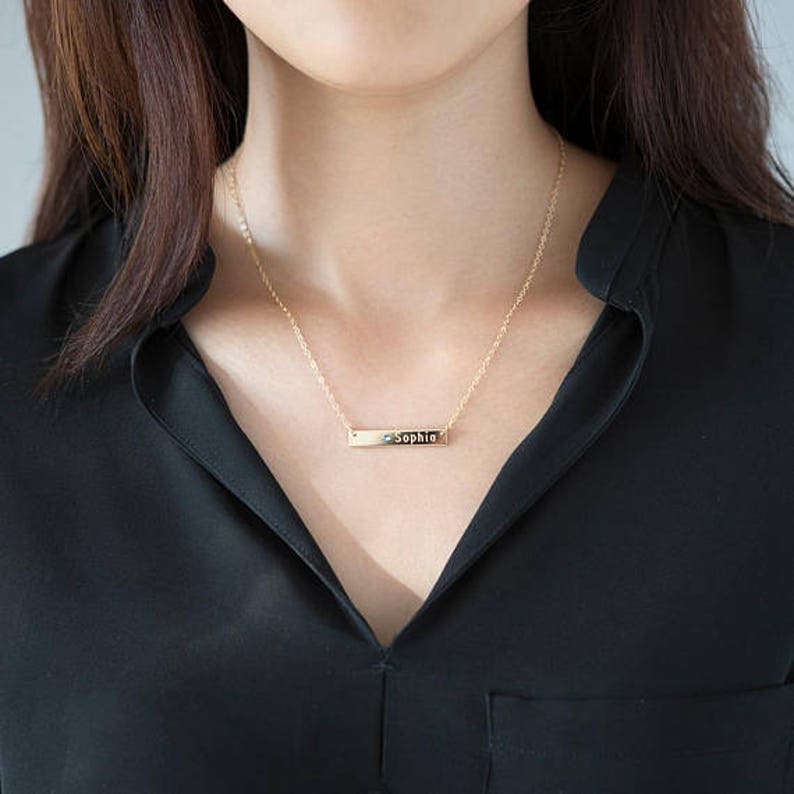Puede incluir: Un collar dorado con un colgante rectangular con el nombre "Sophia" grabado. El collar se lleva sobre una blusa negra.