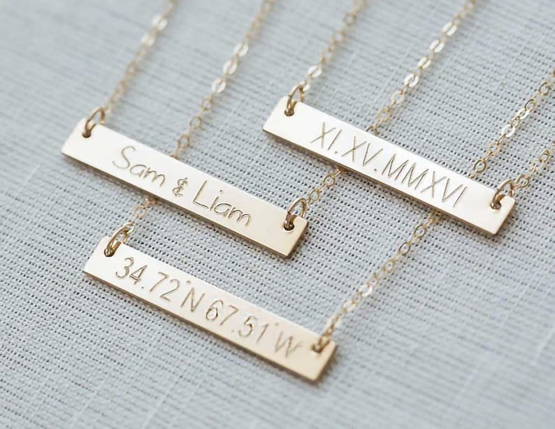 Puede incluir: Tres collares dorados con cadenas delicadas. Cada barra est&aacute; grabada con el texto "Sam & Liam", n&uacute;meros romanos y coordenadas "34.72 N 67.51 W". Los collares se muestran sobre una superficie gris claro.