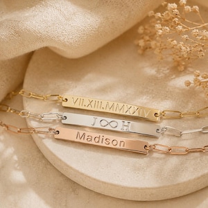 May include: Three personalized bar bracelets in gold, silver, and rose gold. Each bracelet features a rectangular bar with engraved text, including Roman numerals, initials, and a name. The bracelets are displayed on a neutral surface.