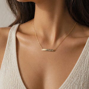 Puede incluir: Un collar dorado con una barra grabada con el nombre "Sophia". El collar se lleva alrededor del cuello y cuelga sobre el pecho. El fondo est&aacute; borroso, centr&aacute;ndose en el collar.