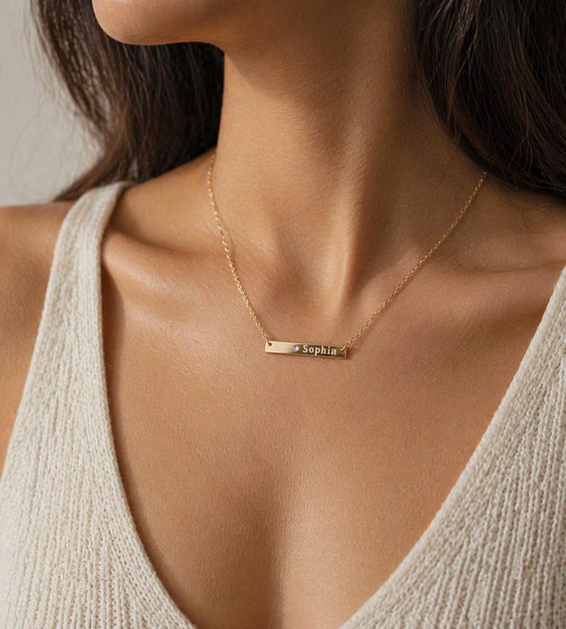 Puede incluir: Un collar dorado con una barra grabada con el nombre "Sophia". El collar se lleva alrededor del cuello y cuelga sobre el pecho. El fondo est&aacute; borroso, centr&aacute;ndose en el collar.