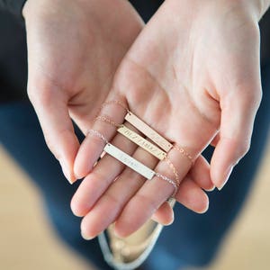 Puede incluir: Tres collares personalizados con delicadas cadenas. Las barras son doradas y plateadas, cada una grabada con una palabra o nombre: "Loved", "GGE" y "VIIZ/IAIA". Los collares se sostienen en manos abiertas sobre un fondo borroso.