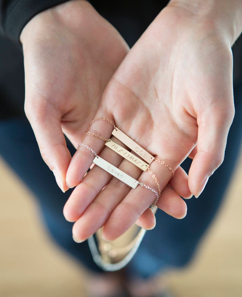 Puede incluir: Tres collares personalizados con delicadas cadenas. Las barras son doradas y plateadas, cada una grabada con una palabra o nombre: "Loved", "GGE" y "VIIZ/IAIA". Los collares se sostienen en manos abiertas sobre un fondo borroso.