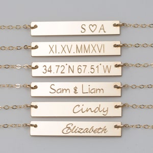Puede incluir: Collares dorados con barras grabadas con texto variado. El texto incluye iniciales, fechas en n&uacute;meros romanos, coordenadas y nombres como "Sam & Liam" y "Cindy". Cada barra est&aacute; conectada a una delicada cadena.