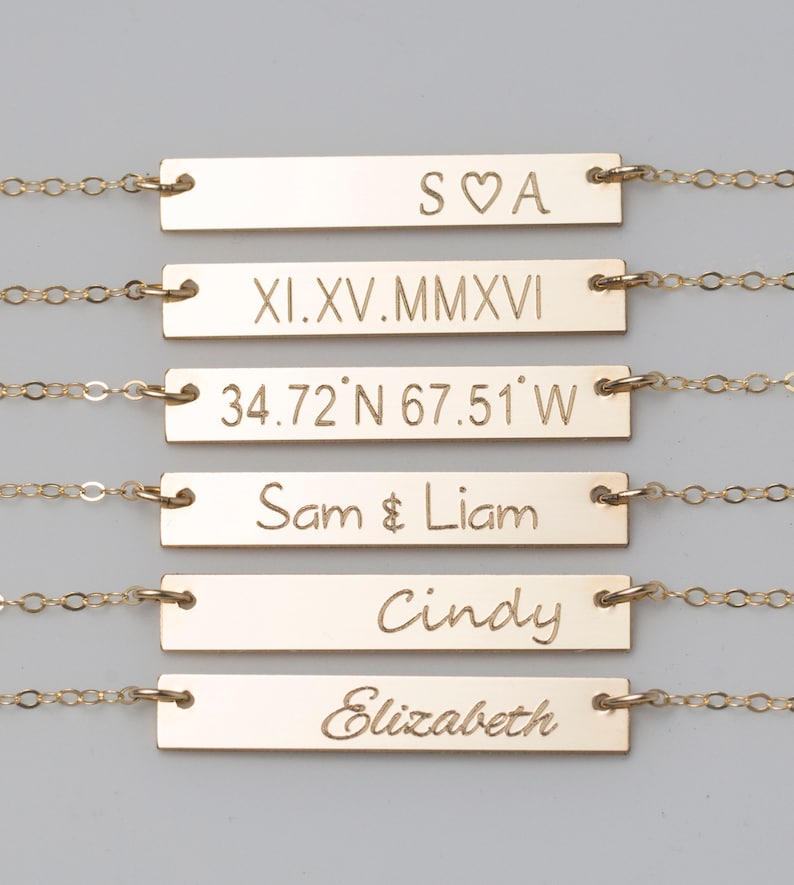 Puede incluir: Collares dorados con barras grabadas con texto variado. El texto incluye iniciales, fechas en n&uacute;meros romanos, coordenadas y nombres como "Sam & Liam" y "Cindy". Cada barra est&aacute; conectada a una delicada cadena.