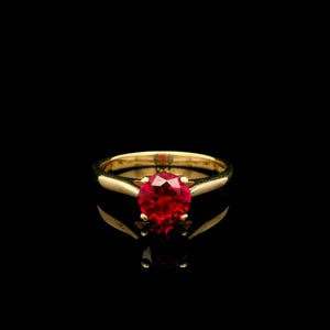 May include: A gold ring featuring a round, faceted, vibrant red gemstone set in a four-prong mounting. The band is smooth and polished, showcasing a classic design with a reflective surface that highlights the deep crimson color of the stone.
