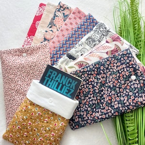 Peut inclure: Plusieurs pochettes de livres en tissu avec divers motifs floraux et imprimés. L'une d'elles contient un livre avec le titre "FRANCK THILLIEZ". Les pochettes sont dans des tons de rose, bleu, jaune et rouge, avec des fermetures à boutons blancs.