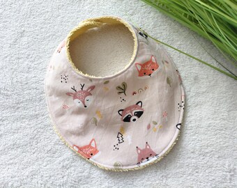 May include: A round baby bib featuring a light pink background with a repeating pattern of woodland animals including foxes, deer, and raccoons, accented by small trees and dots. The bib has a yellow trim and a soft, textured underside.
