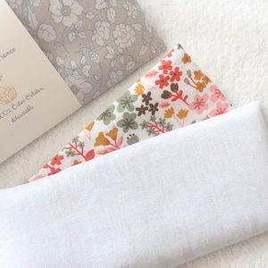 May include: Three rectangular eye pillows with fabric covers. One has a grey floral pattern, one features a multi-colored floral print with pink, orange, and green, and one is plain white. A paper label with text is wrapped around the grey pillow.