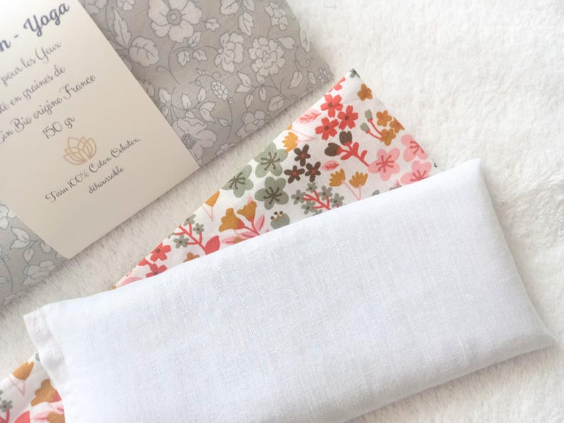 May include: Three rectangular eye pillows with fabric covers. One has a grey floral pattern, one features a multi-colored floral print with pink, orange, and green, and one is plain white. A paper label with text is wrapped around the grey pillow.