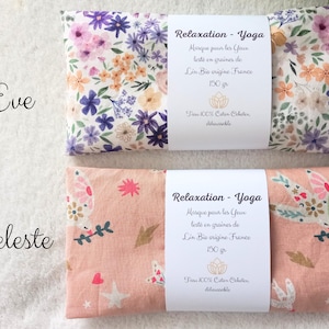 May include: Two rectangular eye pillows labeled Eve and Celeste. The Eve pillow features a white fabric with purple, pink, and orange floral patterns. The Celeste pillow is peach-colored with white bird, star, and heart motifs. Both have white paper bands.
