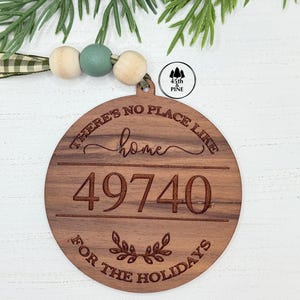 Peut inclure: Ornement de Noël rond en bois avec l'inscription "THERE'S NO PLACE LIKE HOME" et "49740" gravée. L'ornement est orné d'un ruban décoratif et de perles en bois. L'ornement porte également l'inscription "FOR THE HOLIDAYS".