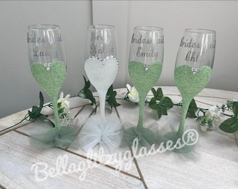May include: Four champagne flutes decorated with green glitter and personalized with the text "bridesmaid" followed by a name. The flutes are adorned with tulle.
