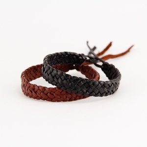 May include: Two braided leather bracelets, one brown and one black, are stacked on top of each other. The bracelets are on a white background.