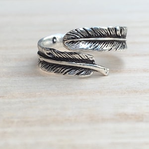 May include: A silver adjustable ring featuring a feather design. The ring is open on one side and can be adjusted to fit different finger sizes.