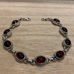 Könnte beinhalten: Ein silbernes Armband mit ovalen, tiefroten Edelsteinen. Das Armband hat ein verziertes Design mit einem Verschluss. Die Edelsteine sind in einer dekorativen Silberfassung gefasst, was einen klassischen und eleganten Look erzeugt.