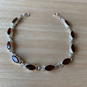 Op de afbeelding: Een zilveren armband met donkerrode edelstenen in een marquise-vorm. De armband heeft een karabijn sluiting.