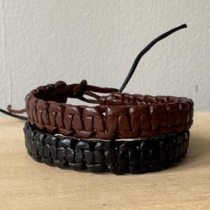 Può includere: Due braccialetti in pelle intrecciata, uno marrone e uno nero. I braccialetti sono impilati uno sopra l'altro.