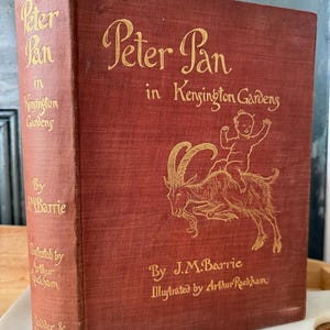 Pode incluir: Um livro vermelho com letras douradas intitulado "Peter Pan nos Jardins de Kensington" de J.M. Barrie, ilustrado por Arthur Rackham. A capa apresenta um desenho de um bebê montando uma cabra.