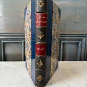 May include: A vintage hardcover book with a blue and gold patterned cover. The spine is black with gold lettering that reads "IZINYANGA ZOKUBULA" and "CALLAWAY". The book was published in 1870.