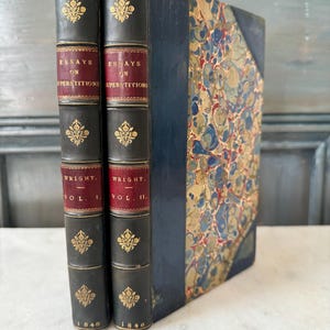 Puede incluir: Dos libros antiguos encuadernados en cuero marrón oscuro con letras doradas. Los libros se titulan "Ensayos sobre supersticiones" y son los volúmenes I y II. Los libros fueron publicados por Wright y datan de 1846.