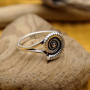 Könnte beinhalten: Ein silberner Ring mit einem Spiraldesign, der auf einem Stück Treibholz liegt. Der Ring befindet sich auf einem gelben Stoffhintergrund mit Weizenähren.