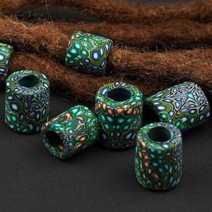 Può includere: Una collezione di perline cilindriche per dreadlock. Le perline sono principalmente verde acqua con un motivo bianco, arancione e giallo. Sono disposte su una superficie scura con extensioni di dreadlock marroni.