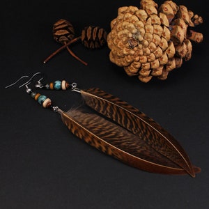 Puede incluir: Un par de pendientes largos de plumas con plumas marrones y negras, cuentas turquesa y marrones y ganchos plateados. Los pendientes se muestran sobre una superficie negra con piñas.