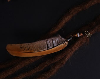 May include: A brown feather with white markings is attached to a brown dreadlock with a small metal charm and brown beads. The feather is lying on a black surface with three brown pine cones.