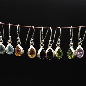 Könnte beinhalten: Eine Auswahl an silbernen Ohrhängern mit tropfenförmigen Edelsteinen in verschiedenen Farben, darunter Blau, Gelb, Orange, Schwarz, Grün und Lila. Die Ohrringe hängen an einem braunen Faden vor einem schwarzen Hintergrund.