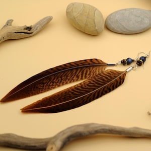 Op de afbeelding: Een paar lange oorhangers met bruine en zwarte veren en blauwe en gouden kralen.