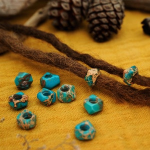 May include: Turquoise blue and brown gemstone beads for dreadlocks. The beads are hexagonal in shape and have a mottled pattern. The beads are strung on two brown dreadlocks.