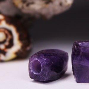 Può includere: Due perline di pietra preziosa di ametista viola, una è quadrata e l'altra rettangolare. Entrambe le perline hanno un foro praticato al centro.