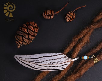 May include: A white and brown feather with black stripes is attached to a brown dreadlock with a small metal bead and a turquoise bead. The feather is on a black surface with brown pine cones.