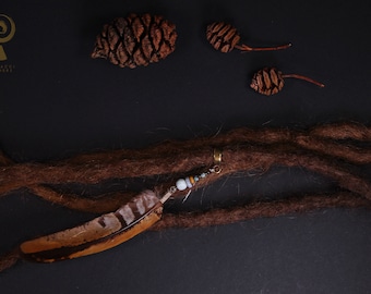 May include: A brown feather hair tie with beads and a gold ring. The hair tie is on a brown dreadlock. There are three pine cones on a black background.
