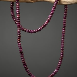 Op de afbeelding: Een lange, gelaagde ketting bestaande uit kleine, ronde, dieprode kralen. De ketting is over een houten tak gedrapeerd tegen een donkergrijze achtergrond. De kralen hebben een gepolijste uitstraling en reflecteren licht.