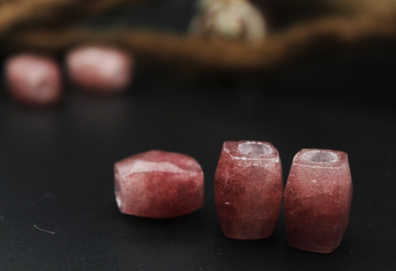 May include: Three pink and white gemstone beads with a faceted, rounded shape. The beads are arranged in a row on a black surface.