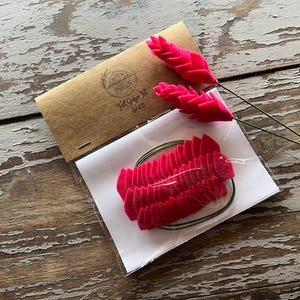 May include: A craft kit featuring felt ginger flowers in bright pink. The kit includes pre-cut felt pieces, wire stems, and a kraft paper package with the text "Felt Ginger Kit". The items are arranged on a distressed wooden surface.