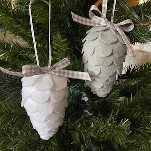 May include: Two felt pinecone ornaments hanging on a Christmas tree. One is white, the other is gray. Both have a ribbon bow at the top with a gingham pattern. The ornaments are made of layered felt pieces.