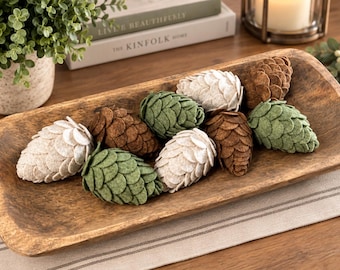 Puede incluir: Una colección de piñas de fieltro en tonos crema, verde oliva y marrón, dispuestas dentro de un cuenco de madera alargado y rectangular tallado a mano. Las piñas de fieltro presentan escamas superpuestas en forma de pétalos.