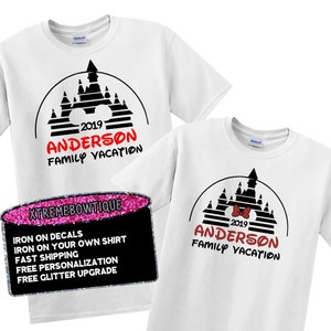 May include: Two white t-shirts with a black castle design, the text "2019 ANDERSON FAMILY VACATION" in red and black. One shirt has a red bow. A sign reads "IRON ON DECALS" and "FREE GLITTER UPGRADE".