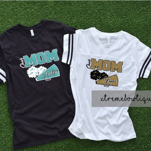 以下が含まれることがあります： 黒と白のストライプのチアリーダーママのTシャツ2枚。1枚は黒地にターコイズ色のグリッター文字と、ターコイズ色のメガホンに「Ashley」の文字が書かれています。もう1枚は白地にゴールドのグリッター文字と、ゴールドのメガホンに「Lions」の文字が書かれています。