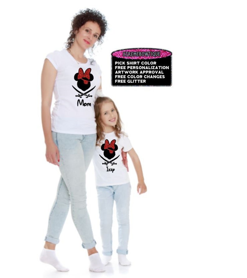 May include: Two white t-shirts with a Minnie Mouse pirate design. One shirt says "Mom" and the other says "Izzp". The design includes a red bow, crossed swords, and a black pirate hat. The shirts are worn with light blue jeans.