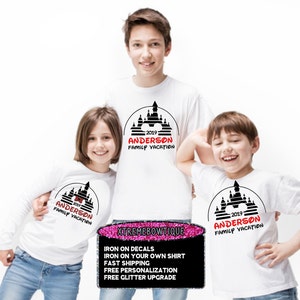May include: Three white t-shirts with a black and red castle design and the text "2019 ANDERSON FAMILY VACATION." The shirts are worn by three children. A sign at the bottom advertises iron-on decals and personalization.