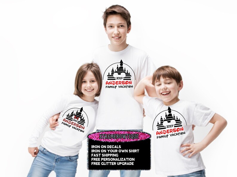 May include: Three white t-shirts with a black and red castle design and the text "2019 ANDERSON FAMILY VACATION." The shirts are worn by three children. A sign at the bottom advertises iron-on decals and personalization.