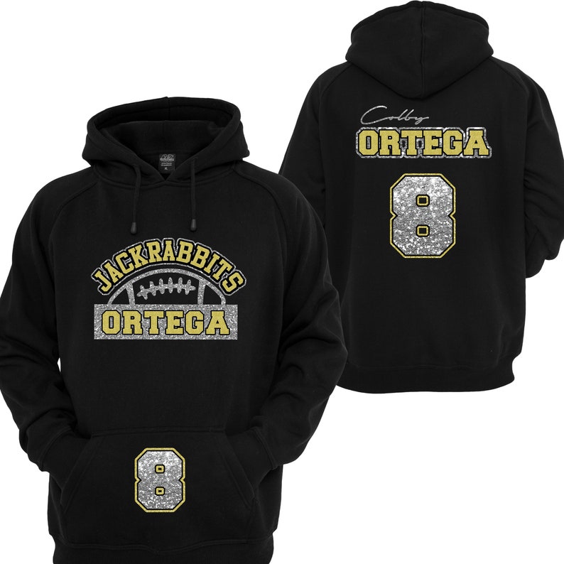 May include: Black hoodie with a silver and gold glitter design. The front of the hoodie has the text "JACKRABBITS" in a semi-circle above a football and the text "ORTEGA". The back of the hoodie has the text "Colby ORTEGA" and the number "8" in silver and gold glitter.